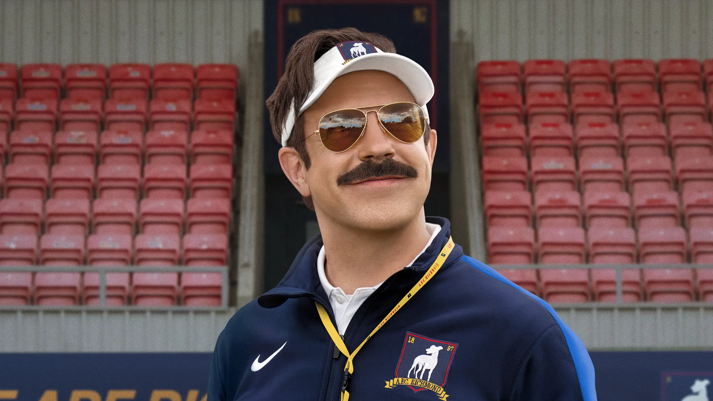 Ted Lasso napszemüvegben, egy stadion lelátója előtt az Apple TV hivatalos műsorképén.