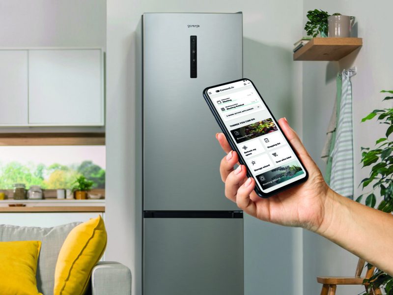 Gorenje hűtő előtt kézben tartott telefon, kijelzőjén a ConnectLife alkalmazás készülék- és receptkezelő felületével.